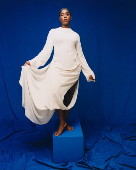 Portrait of Lashana Lynch in long white dress against blue background stand on blue cube