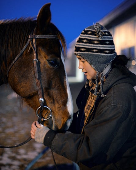 Lily Gladstone in a ear-flapped hat, talking to a horse