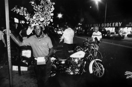 A black and white photo of war veteran Floyd wearing vast foliage on his head, holding a box and standing next to men and a motorcycle, outside a deli.