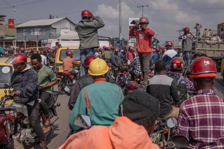 Men on motorbikes block a road, with some standing on their saddles making gun gestures at their heads while covering their mouths.