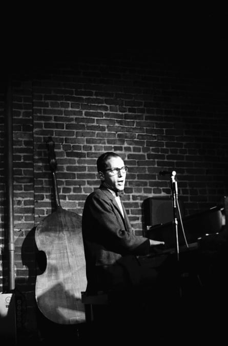 Black and white shows Lehrer singing and playing piano in a spotlight on stage, with a brick wall behind him and a double bass facing the wall.