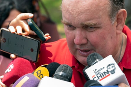 A man wearing a red shirt speaks in front of several news microphones.