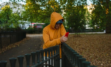 Teenage boy in yellow hooded sweater, using his cell phone leaning against a fence in a London park.