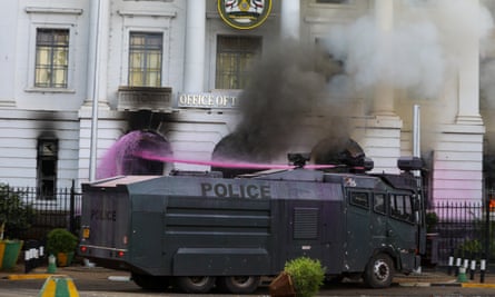 Pink water shoots from a cannon on top of a large black police vehicle towards a grand building from which smoke is billowing