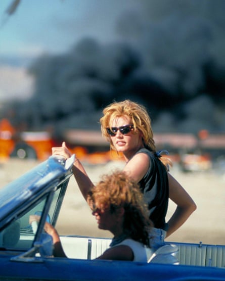 Geena Davis stands in a car, with Susan Sarandon sitting in the driving seat. In the background is billowing black smoke