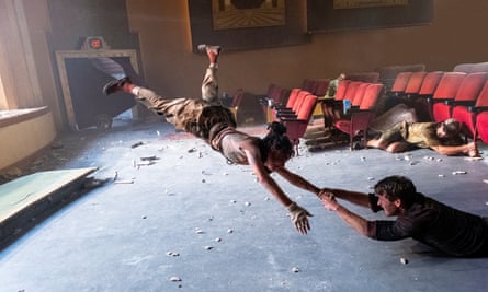 With her legs elevated into the air, Sasha Lane, in a T-shirt and slightly baggy trousers, holds onto Powell’s hand as he lies on the floor with cinema seats behind them while an audience member in a baseball cap shouts and holds onto a seat