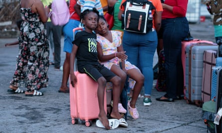 Two girls sit on a pink suitcase as adults with bags and suitcases stand nearby