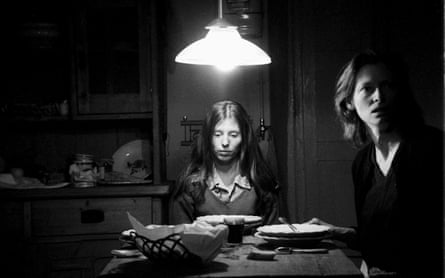 Two woman sit at a table dimly illuminated by an overhead lamp - one woman on the right looking off to the right