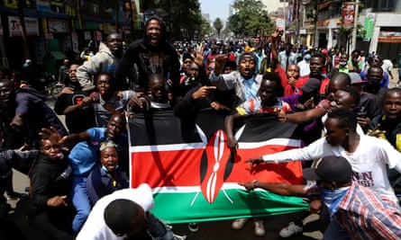 Demonstrators carry a Kenyan flag during a protest in Nairobi, Tuesday 23 July 2024: young men and women stand around and behind the flag shouting and chanting and gesticulating; the crowd stretches into the distance down the street