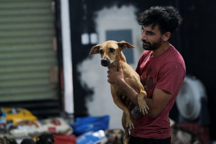 A young man with black hair and heard and a red T-shirt holds a short-haired brown dog, smiling.