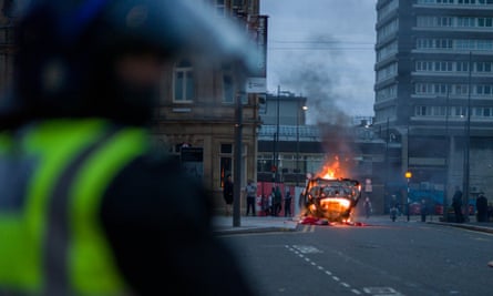 A police car on fire in a street with people standing nearby.