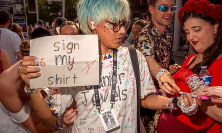 A fan with turquoise hair holds a sign reading ‘Sign my shirt’ holds a pen out to a woman. His white T-shirt is already covered in writing