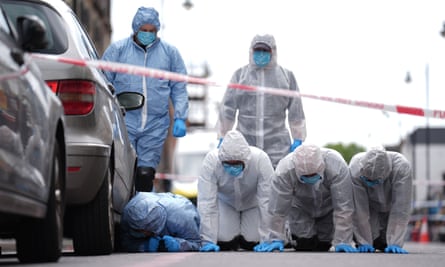 Four investigators in plastic suits, gloves and hats on their hands and knees on the street next to cars and crime scene tape stretched across