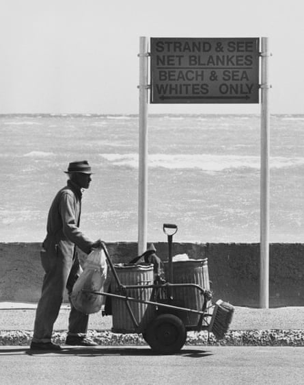 A man in a trilby and pushing a dustcart walks past a beachside sign that reads: ‘Strand & see net blankes beach’ alongside the English: ‘Beach & sea whites only’