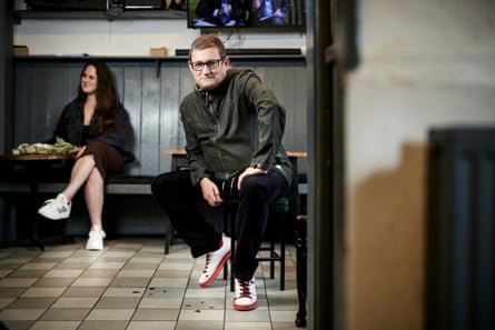 Heaton sits at a seat in a pub with a tiled floor and wooden panelled wall. A woman sits on a bench in the background