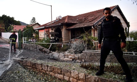 An Israeli police officer stands by as a firefighter sprays water from a hose by a house