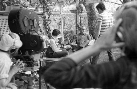 A black-and-white photograph of Al Pacino and Marlon Brando sitting in garden armchairs by a small table under a pergola, with a man with a large film camera and another with headphones in the foreground, and a third man standing by the actors