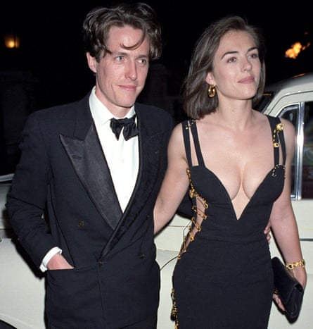 Hugh Grant, in a tuxedo, with his arm around Elizabeth Hurley in her low-cut dress with safety pins down the sides