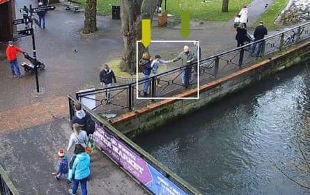 blurred CCTV image showing three people at the edge of a pond; a man is passing a piece of bread to a boy to feed the ducks