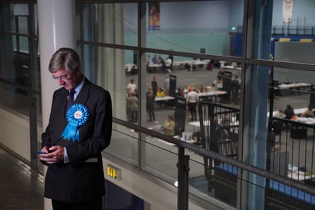 Rees-Mogg in suit and blue rosette, looking at his phone while in the distance the counting of votes that will depose him goes on.