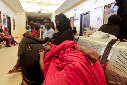 A woman lies on a bench in a hospital waiting area with her head on her relative’s lap