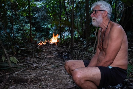 A man with short white hair and a short white beard sits topless in the jungle, with a campfire in the background.