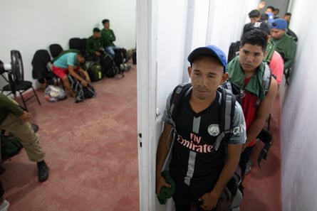 A line of young men wait in a corridor wearing backpacks and holding or wearing dark green t-shirts. Through an open door we can see more young men wearing the shirts sitting on chairs with their packs at their feet.