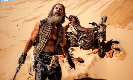 man wearing vest holds weapon and looks up as bike sits behind him in sand