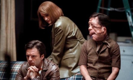 man wearing brown shirt looks to his right as another man sits on plaid couch with woman wearing tan jacket standing behind them