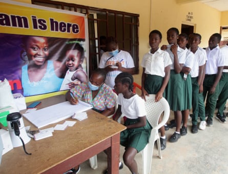 A child receives a vaccine at a school desk, while a row of schoolchildren line up behind her