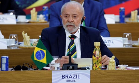 a man in suit and tie seated at a desk bearing a Brazilian flag