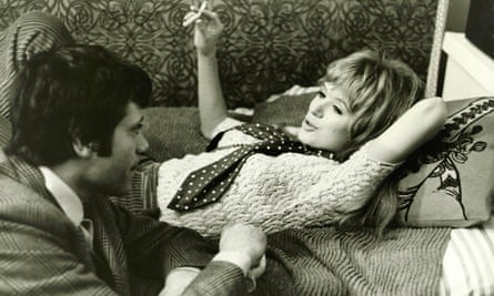 a woman lays on a couch smoking a cigarette as a man crouches next to hear