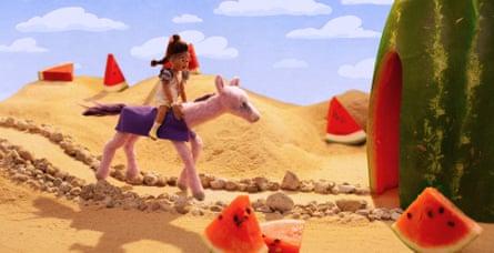 An animated still showing a sandy desert landscape with pieces of real watermelon, and a girl made from felt sitting on a pink horse made from felt, making their way down a pathway towards a building made from a watermelon