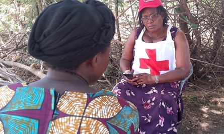 Judith Fukizi, wearing a white bib with a red cross on it, sits opposite a woman in the shade under some trees