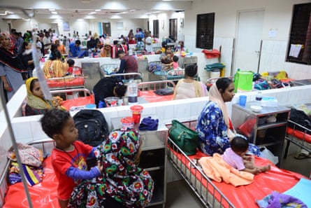 Dozens of people and small children sit on beds in bays in a large medical ward.