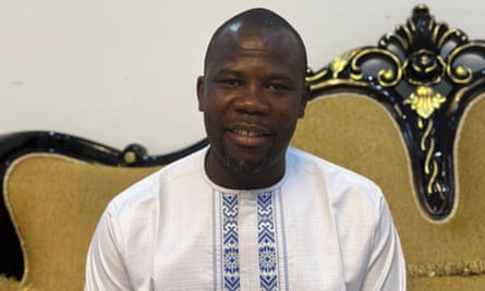 Portrait of Abdoulaye Kourouma: he sits on an ornate couch with black and gold frame and wears a white tunic with blue embroidery down the front