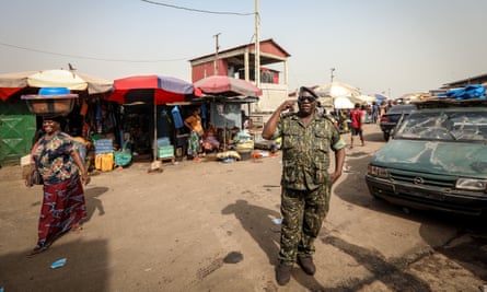 A police officer stands guard: he wears camouflage shirt and trousers, a beret and dark glasses, and stands by a minivan with a broken windscreen. A woman walks behind him carrying a large container on her head and there are stalls under umbrellas in the background