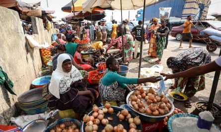 A local market: women sit by a wall under sunshade umbrellas surrounded by large bowls of onions while others sell peppers, aubergines, tomatoes and other goods behind them