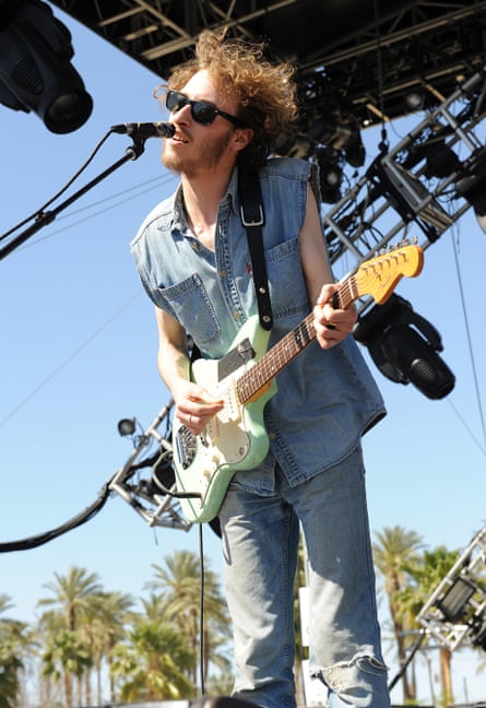 Daniel Blumberg in denim shortsleeved top and jeans playing guitar in shades and with wild curly hair 