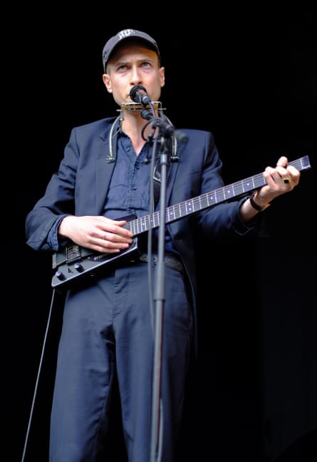 Daniel Blumberg in a blue shirt and suit and blue cap playing guitar and mouth organ