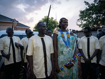 A young woman in traditional clothing and carrying a beaded sceptre stands next to a shorter man wearing a traiditional shirt with