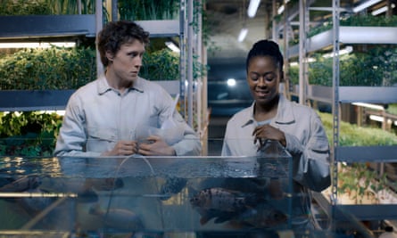 George MacKay and Moses Ingram standing next to pool of water in a lab-like room full of verdant shelves