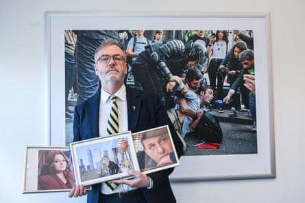 An older man holding photos of three people with a large photo on the wall behind him of riot police manhandling a protester