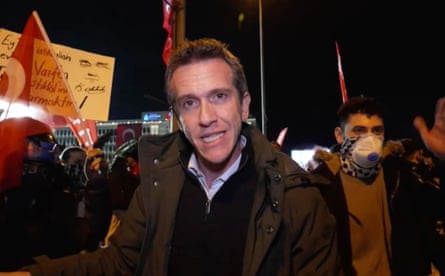A middle-aged man speaks to a camera in a demonstration with flags and placards held by protesters behind