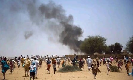 A large crowd of people running across a desert-like landscape as black smoke billows from a compound in the background
